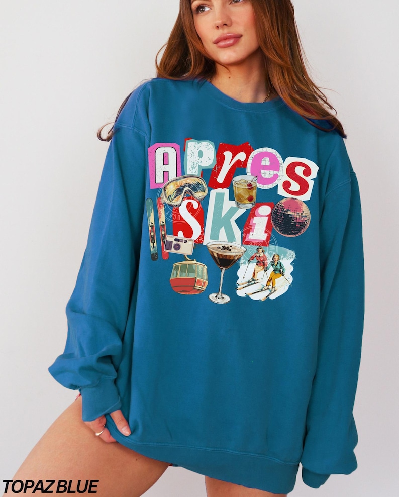 May include: Topaz blue sweatshirt with a colorful graphic design. The design includes the words "Apres Ski" with images of ski goggles, a cocktail, a disco ball, and skiers. The sweatshirt is a crew neck style.