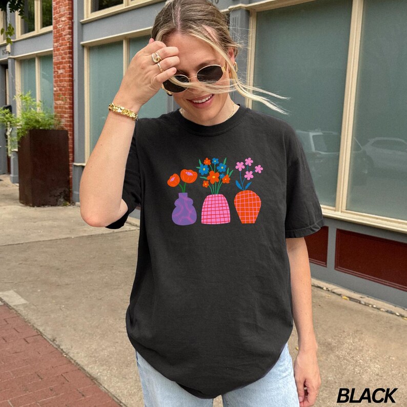 May include: Black t-shirt with a graphic of four colorful vases with flowers. The vases are in a row and the flowers are in different colors and styles. The text "BLACK" is visible in the bottom right corner of the image.