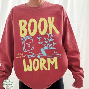 May include: Crimson-colored sweatshirt with a yellow "BOOK WORM" graphic. The design includes a book, glasses, and a potted plant. The text "all day every day" is also included in the design.