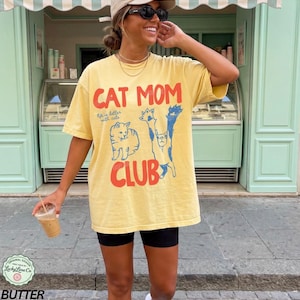 Puede incluir: Camiseta amarilla con el texto "CAT MOM CLUB" en rojo y azul, con ilustraciones de gatos. La camiseta se lleva con pantalones cortos negros. La imagen incluye un logotipo que dice "Original Design Lucky Lime Co" y la palabra "BUTTER".