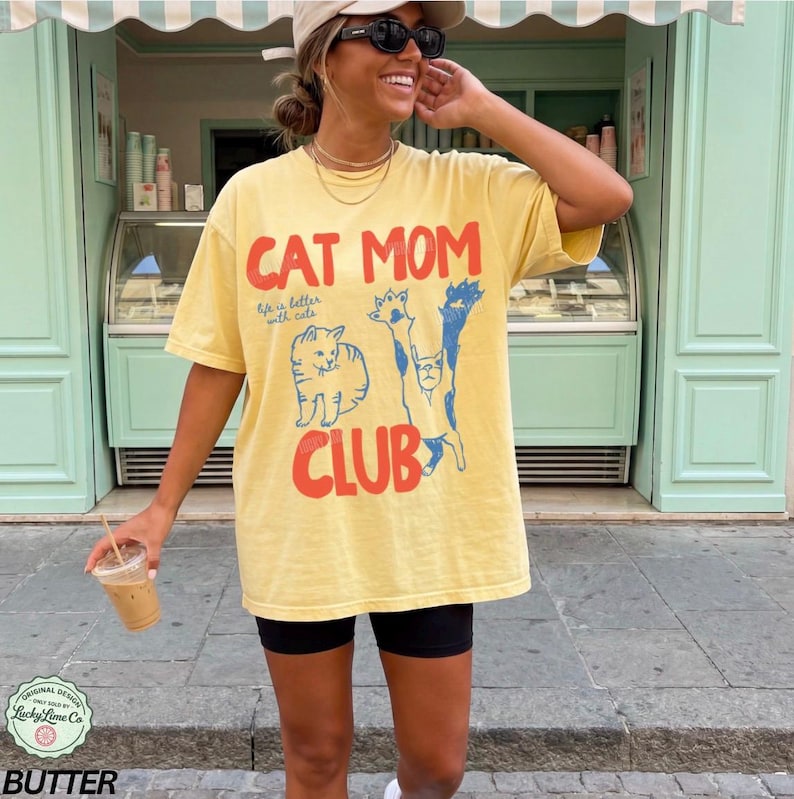 Puede incluir: Camiseta amarilla con el texto "CAT MOM CLUB" en rojo y azul, con ilustraciones de gatos. La camiseta se lleva con pantalones cortos negros. La imagen incluye un logotipo que dice "Original Design Lucky Lime Co" y la palabra "BUTTER".