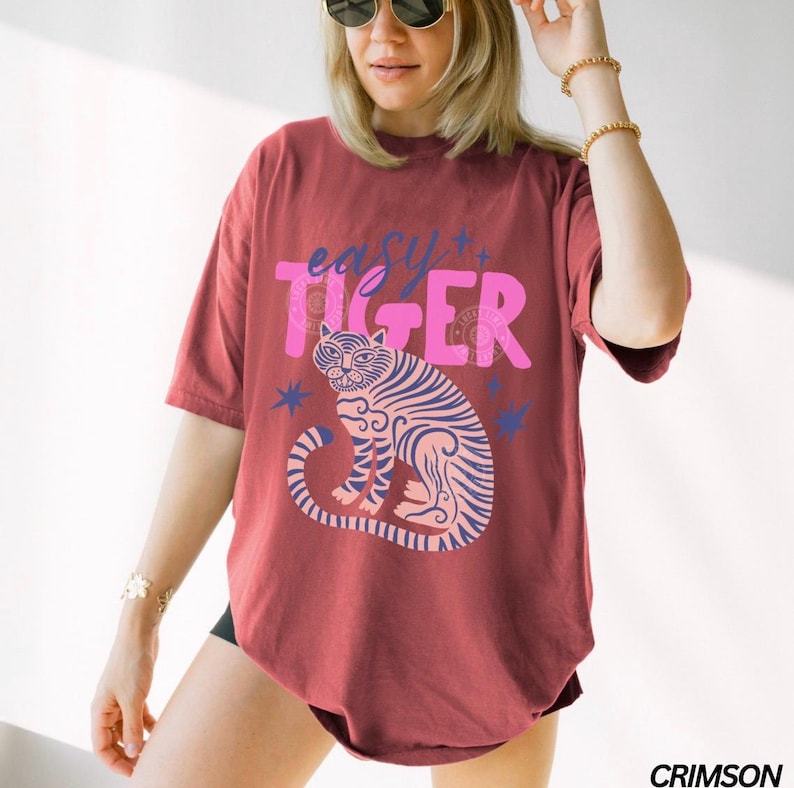 Puede incluir: Camiseta color carmes&iacute; con la frase "easy tiger" en rosa y una ilustraci&oacute;n de tigre rosa y azul. La camiseta tiene mangas cortas y un corte holgado.