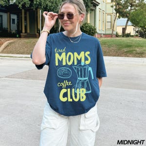 May include: A navy blue t-shirt with the text "tired MOMS coffee CLUB" in yellow and a coffee pot graphic. The shirt is paired with white cargo pants. The image is taken outdoors.