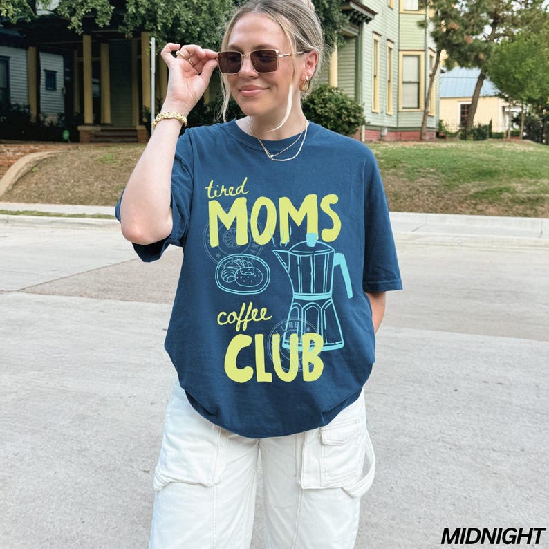 May include: A navy blue t-shirt with the text "tired MOMS coffee CLUB" in yellow and a coffee pot graphic. The shirt is paired with white cargo pants. The image is taken outdoors.