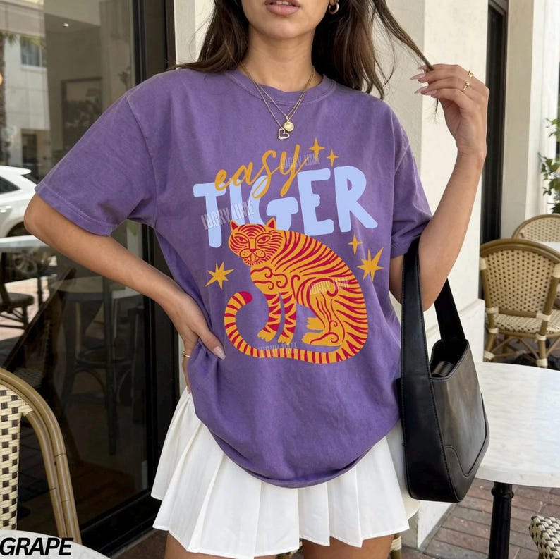 May include: A grape-colored t-shirt with the text "easy tiger" and a tiger graphic in orange and yellow. The shirt is paired with a white pleated skirt and a black shoulder bag. The model is wearing a necklace.