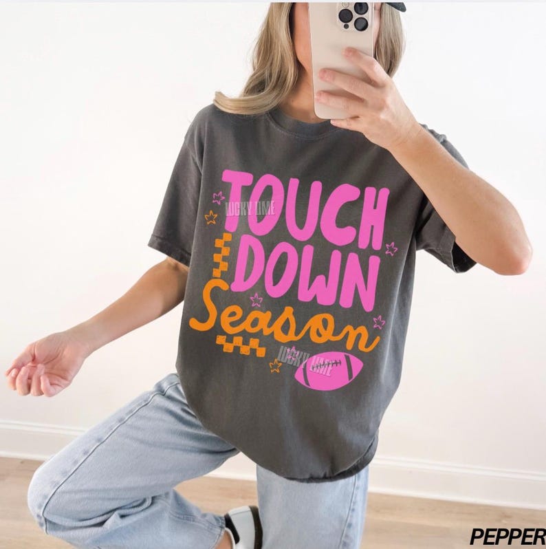May include: Dark grey t-shirt with the words "TOUCH DOWN Season" in pink and orange. The shirt features a football graphic and star accents. The text "LUCKY TIME" is also visible. The shirt is a casual style.