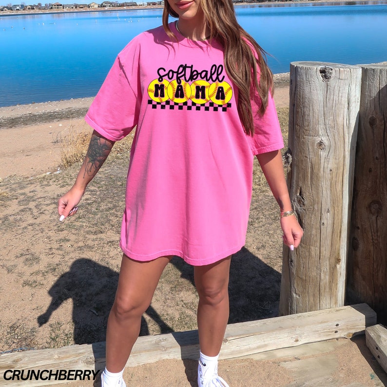 Op de afbeelding: Een roze T-shirt met de tekst "Softball Mama" in zwarte letters en gele softball-afbeeldingen.