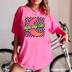 May include: Pink oversized t-shirt with a graphic of a crescent moon and checkered pattern. The text "night shift crew" is written in green and yellow letters.