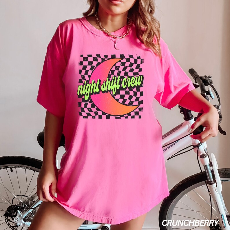 May include: Pink oversized t-shirt with a graphic of a crescent moon and checkered pattern. The text "night shift crew" is written in green and yellow letters.