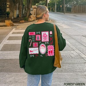 May include: Forest green sweatshirt with a festive design on the back. The design includes Christmas-themed postage stamp graphics with a house, Santa, and candy canes. The text "HO HO HO" is also featured. The sweatshirt is paired with blue jeans.