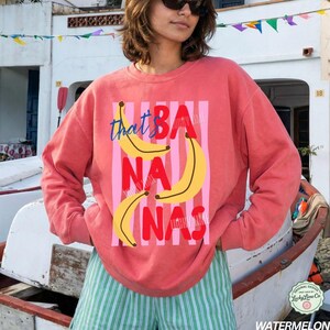 May include: Coral-colored sweatshirt with the text "that's BANANAS" in a playful font, featuring yellow banana illustrations and pink vertical stripes. The sweatshirt has long sleeves and a relaxed fit.