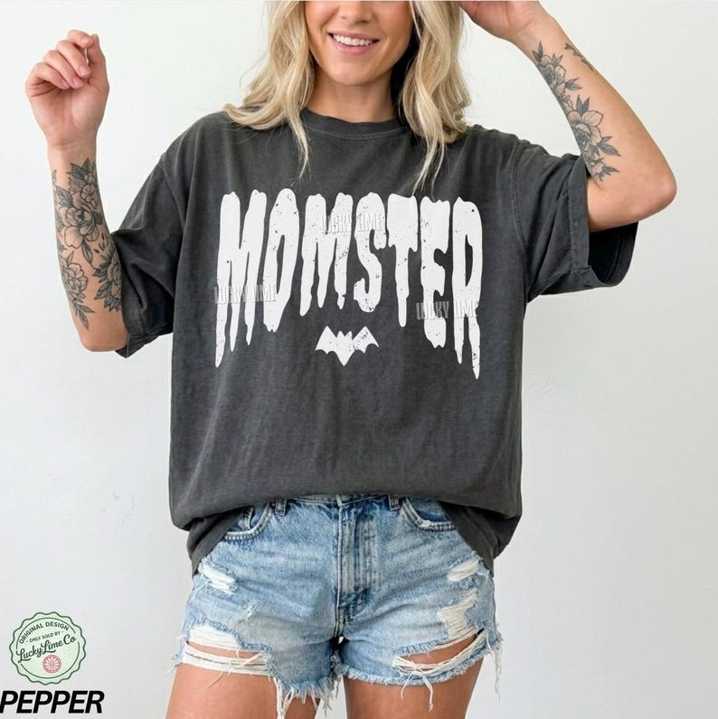May include: Dark gray t-shirt with the word "MOMSTER" in white, a bat graphic, and the text "Lucky Lime Co". The shirt is a relaxed fit and is paired with denim shorts.