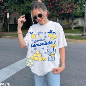 以下が含まれることがあります： 青と黄色の「Homemade Lemonade」グラフィックがプリントされた白いTシャツ。デザインにはレモン、花、レモネードのグラスが含まれています。「fresh squeezed」の文字も表示されています。