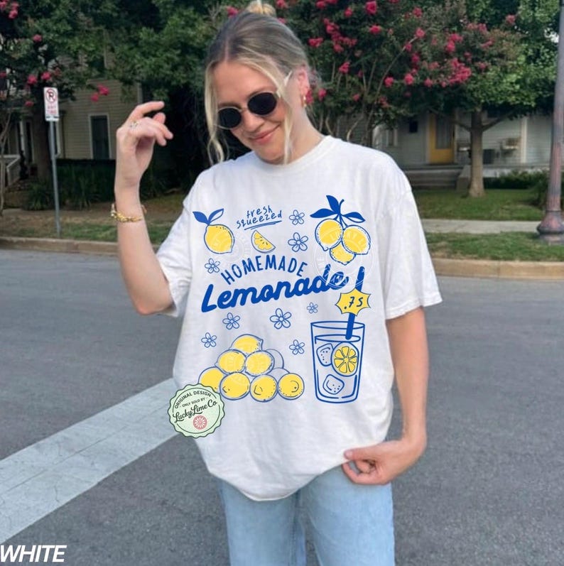 以下が含まれることがあります： 青と黄色の「Homemade Lemonade」グラフィックがプリントされた白いTシャツ。デザインにはレモン、花、レモネードのグラスが含まれています。「fresh squeezed」の文字も表示されています。