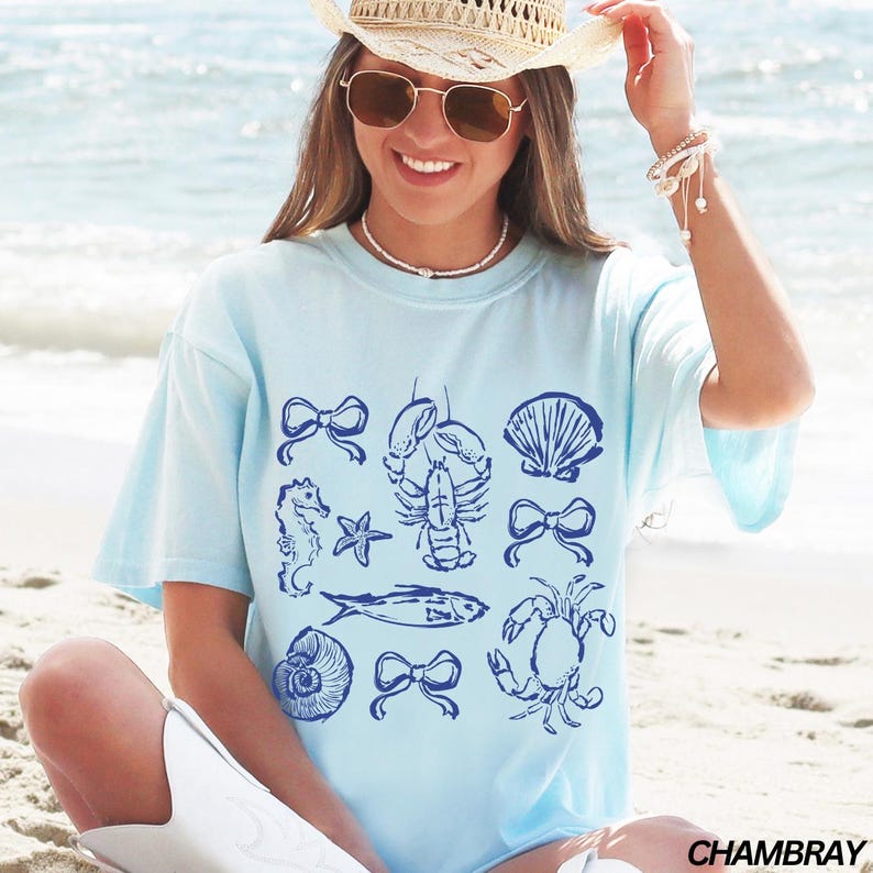 May include: A light blue t-shirt with a blue graphic print of various sea creatures, including a lobster, a seahorse, a crab, a fish, and a seashell. The graphic also includes bows and stars. The shirt has a Chambray label.