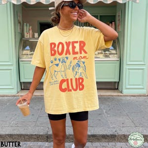 May include: A pale yellow short-sleeve t-shirt featuring a graphic print with the red text BOXER and CLUB, and the blue text mom. The design includes two blue line-art illustrations of boxer dogs, paw prints, and the text Life is better with boxers.