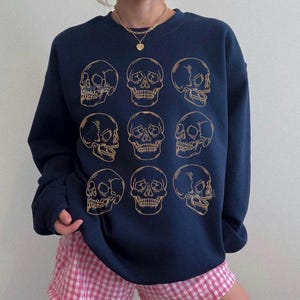 May include: A navy blue crewneck sweatshirt featuring a grid of nine gold-colored line art skull illustrations arranged in three rows of three. The skulls are depicted from various angles, including front-facing and profile views, printed on the chest.
