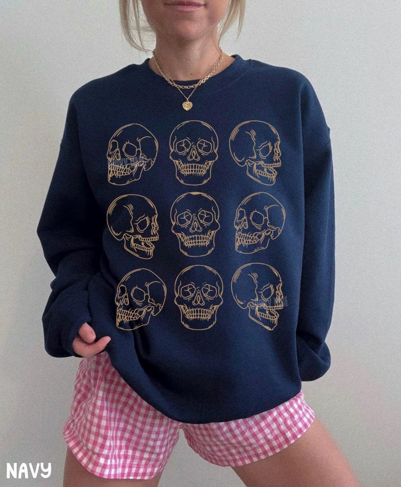 May include: A navy blue crewneck sweatshirt featuring a grid of nine gold-colored line art skull illustrations arranged in three rows of three. The skulls are depicted from various angles, including front-facing and profile views, printed on the chest.