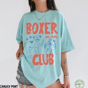 May include: A light mint green t-shirt featuring the red text "BOXER mom CLUB" and blue line art illustrations of two boxer dogs. The design includes small paw prints and the text "life is better with boxers" in a cursive script font.