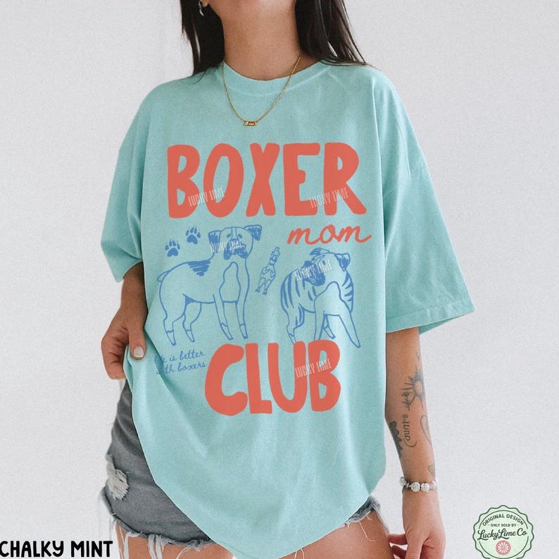 May include: A light mint green t-shirt featuring the red text "BOXER mom CLUB" and blue line art illustrations of two boxer dogs. The design includes small paw prints and the text "life is better with boxers" in a cursive script font.