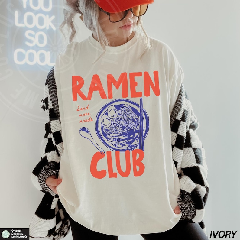 May include: A white long-sleeved shirt with a graphic of a bowl of ramen noodles and the text "Ramen Club" in red. The text "Send more noods" is written in black below the bowl.