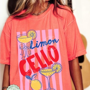May include: Bright salmon-colored t-shirt with a graphic design featuring the words "Limon Cello" in blue and red, along with illustrations of cocktails and lemons. The shirt has a relaxed fit and short sleeves.
