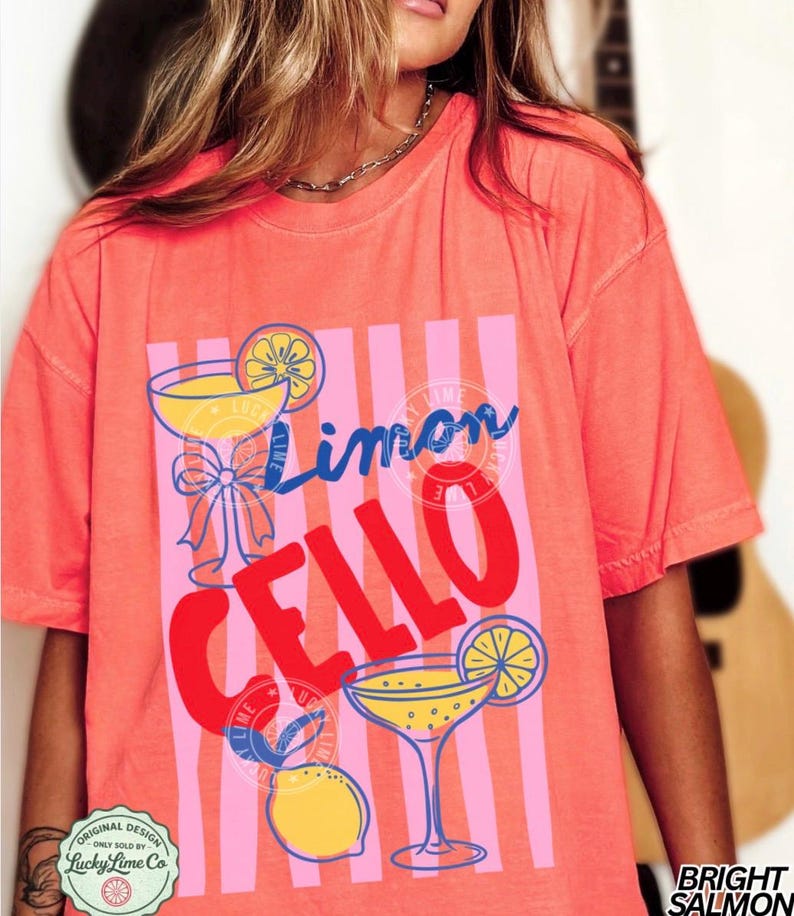 May include: Bright salmon-colored t-shirt with a graphic design featuring the words "Limon Cello" in blue and red, along with illustrations of cocktails and lemons. The shirt has a relaxed fit and short sleeves.