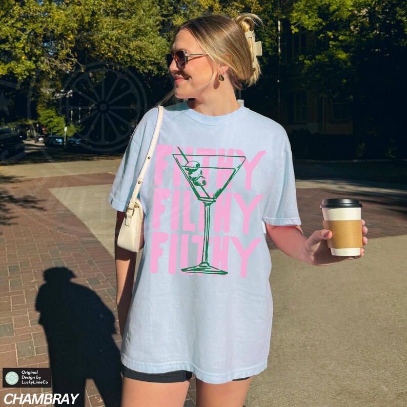 Pode incluir: Uma camiseta azul claro com um gr&aacute;fico rosa de uma ta&ccedil;a de martini com azeitonas. O texto "Filthy Filthy Filthy" est&aacute; impresso na camiseta.