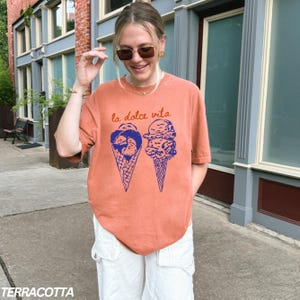 May include: A terracotta orange t-shirt with a graphic of two ice cream cones. The text "la dolce vita" is printed above the ice cream cones.