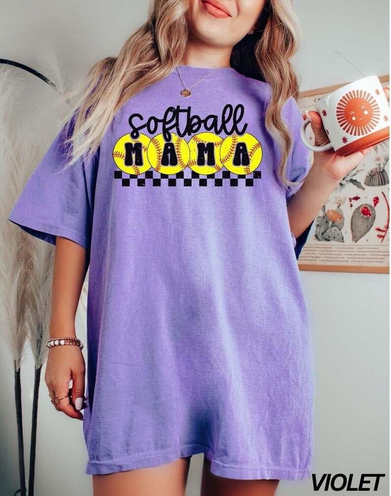 Op de afbeelding: Paars T-shirt met de tekst "Softball Mama" in zwart met gele softball-afbeeldingen en een zwart-wit geruit patroon.