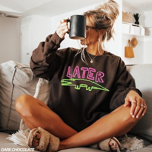 May include: Dark chocolate brown sweatshirt featuring the word "LATER" in pink and a green alligator graphic. A black mug is held up. The sweatshirt has a relaxed fit.