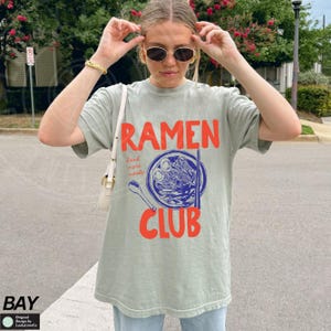 May include: A light green t-shirt with a graphic of a bowl of ramen noodles and the text "Ramen Club" in red and blue. The text "Send more noodles" is written in smaller blue text.