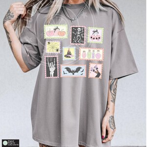 May include: Grey t-shirt featuring Halloween-themed postage stamp designs. The stamps depict pumpkins, skeletons, bats, witch hats, potion bottles, and a skeleton hand. The text "GREY" is at the bottom of the shirt.
