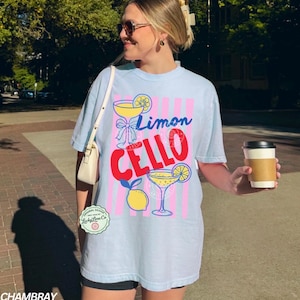 May include: Light blue t-shirt with a "Limon Cello" graphic. The design features a cocktail glass, lemon, and pink and blue stripes. The shirt has a relaxed fit and short sleeves.