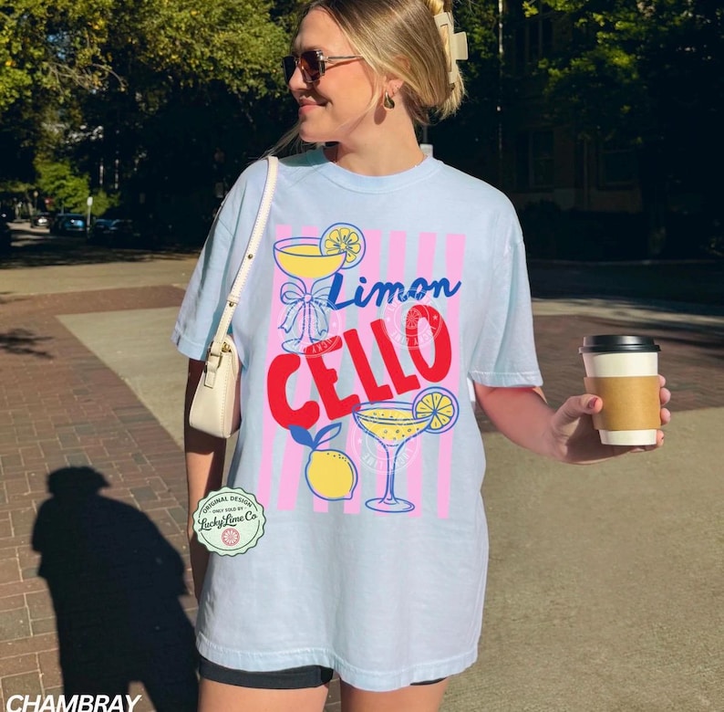 May include: Light blue t-shirt with a "Limon Cello" graphic. The design features a cocktail glass, lemon, and pink and blue stripes. The shirt has a relaxed fit and short sleeves.