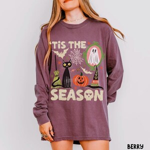Może przedstawiać: Jagodowa koszulka z długim rękawem z nadrukiem graficznym i napisem 'TIS THE SEASON'. Wz&oacute;r przedstawia czarnego kota, dynię, kapelusz czarownicy, miotłę, ducha w zielonej ramce, pajęczyny oraz nietoperze w r&oacute;żnych kolorach.
