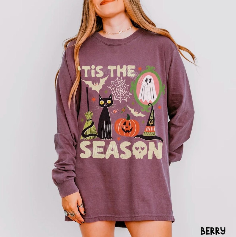 Może przedstawiać: Jagodowa koszulka z długim rękawem z nadrukiem graficznym i napisem 'TIS THE SEASON'. Wz&oacute;r przedstawia czarnego kota, dynię, kapelusz czarownicy, miotłę, ducha w zielonej ramce, pajęczyny oraz nietoperze w r&oacute;żnych kolorach.