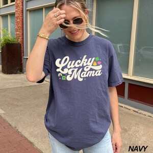 Peut inclure: Un t-shirt bleu marine avec un motif blanc r&eacute;tro qui dit "Lucky Mama" avec un arc-en-ciel, un tr&egrave;fle et un c&oelig;ur.