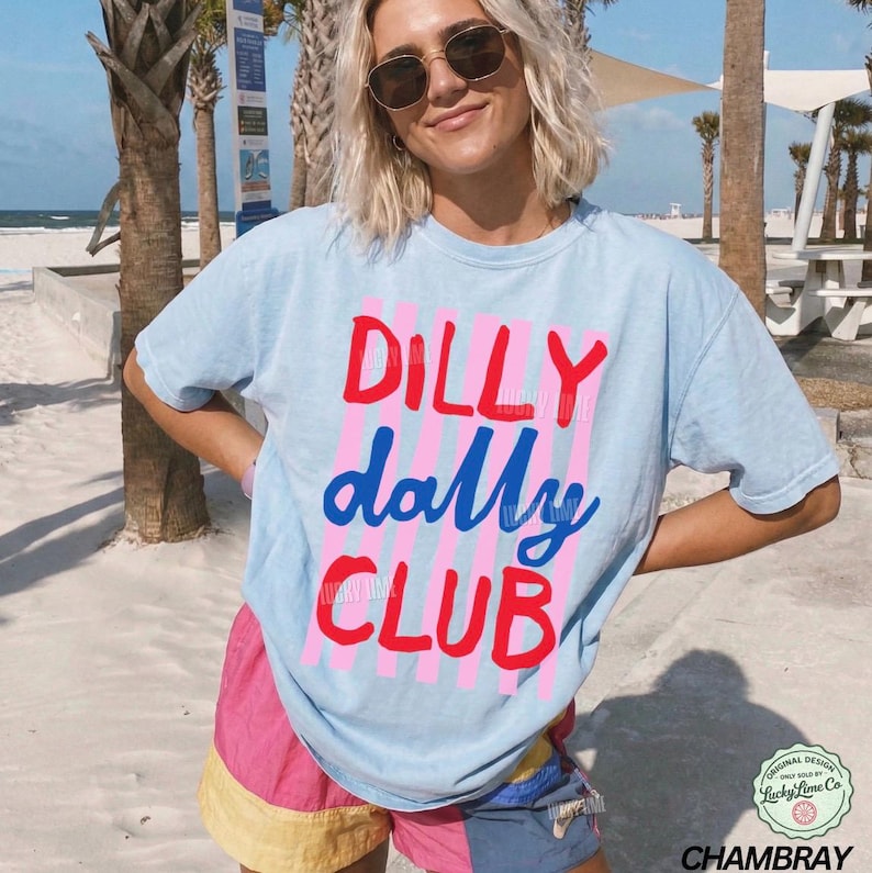 May include: Light blue t-shirt with the words "DILLY dally CLUB" in red and blue lettering. The shirt has pink vertical stripes behind the words. The shirt is part of the Chambray collection.