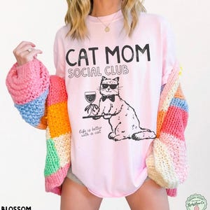 K&ouml;nnte beinhalten: Ein hellrosa T-Shirt mit dem schwarzen Text CAT MOM SOCIAL CLUB und einer Grafik einer Katze mit Sonnenbrille und Fliege, die ein Weinglas auf einem Tablett h&auml;lt. Unter der Katze steht der Text Life is better with a cat in schwarzer Schrift.