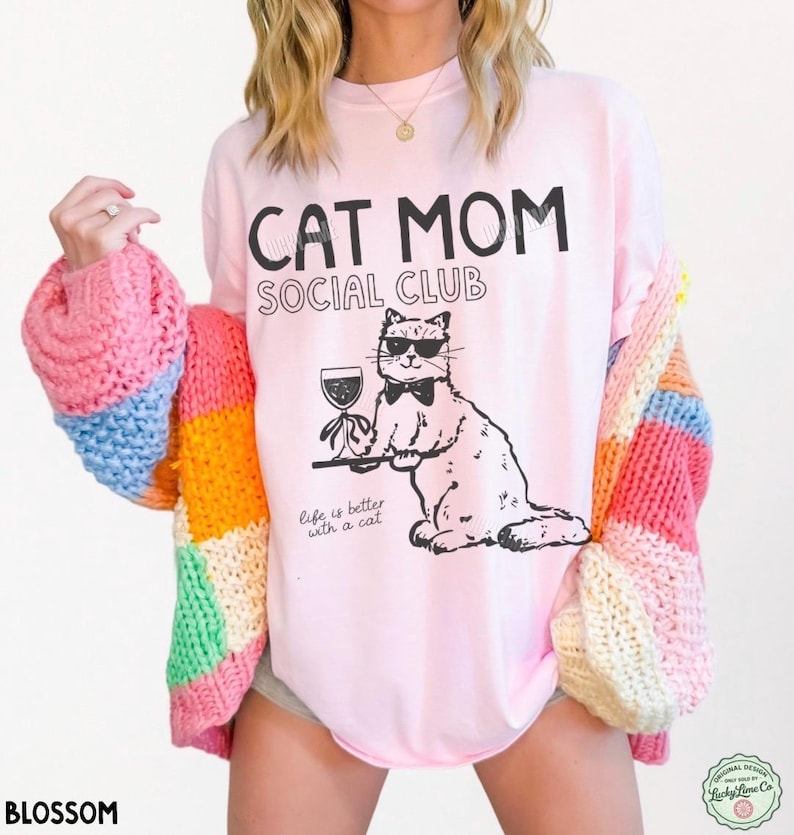 K&ouml;nnte beinhalten: Ein hellrosa T-Shirt mit dem schwarzen Text CAT MOM SOCIAL CLUB und einer Grafik einer Katze mit Sonnenbrille und Fliege, die ein Weinglas auf einem Tablett h&auml;lt. Unter der Katze steht der Text Life is better with a cat in schwarzer Schrift.
