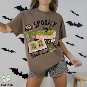 Op de afbeelding: Espresso-kleurig T-shirt met een afbeelding met de tekst "Spooky Book Club". Het ontwerp toont een spin, boeken en rozen. Het shirt wordt gecombineerd met een zwart-witte geruite short. Vleermuizen versieren de achtergrond.