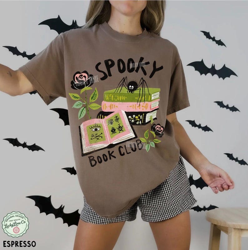 Op de afbeelding: Espresso-kleurig T-shirt met een afbeelding met de tekst "Spooky Book Club". Het ontwerp toont een spin, boeken en rozen. Het shirt wordt gecombineerd met een zwart-witte geruite short. Vleermuizen versieren de achtergrond.