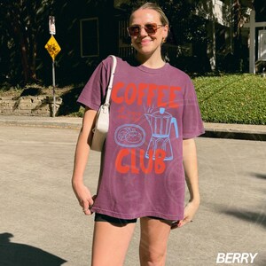 Op de afbeelding: Een paars T-shirt met de tekst "Coffee Club" en een afbeelding van een koffiepot en een bord met gebak.