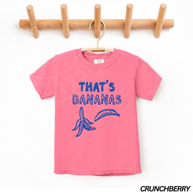 Op de afbeelding: Roze T-shirt met korte mouwen met een blauwe afbeelding van twee bananen en de tekst "That's Bananas".