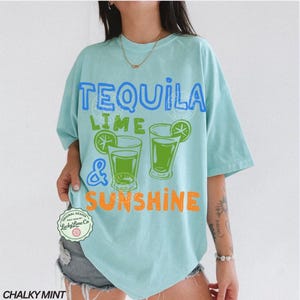 May include: A turquoise t-shirt with the text "TEQUILA LIME & SUNSHINE" in blue, green, and orange. The design includes two green cocktail glasses with lime wedges. The shirt is a loose fit with short sleeves.