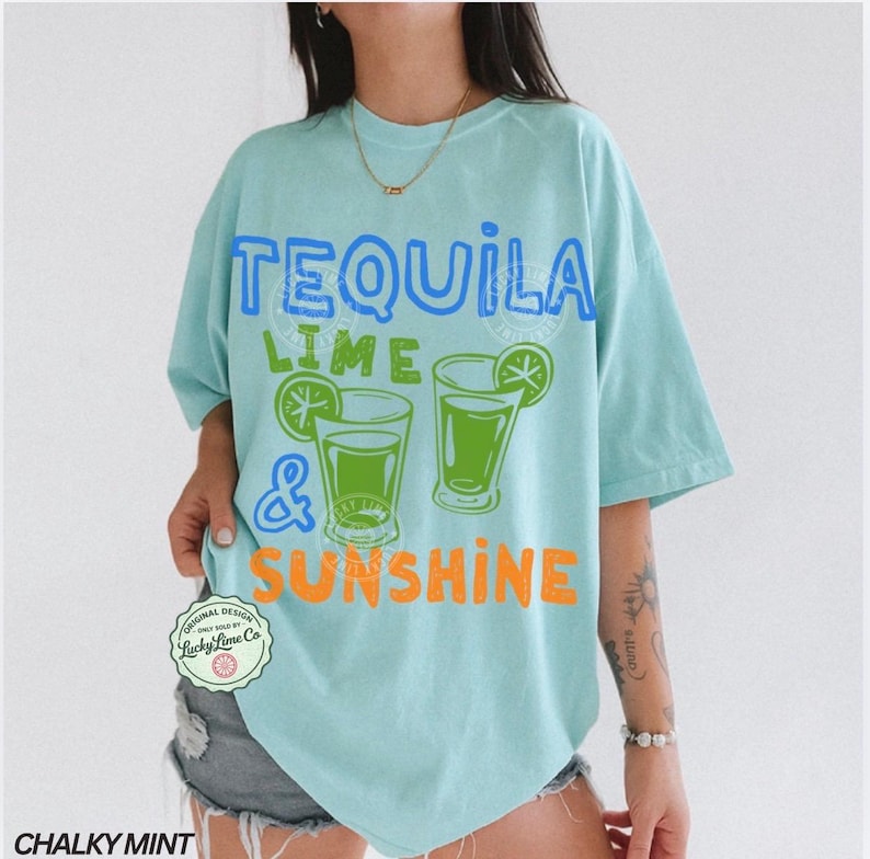May include: A turquoise t-shirt with the text "TEQUILA LIME & SUNSHINE" in blue, green, and orange. The design includes two green cocktail glasses with lime wedges. The shirt is a loose fit with short sleeves.