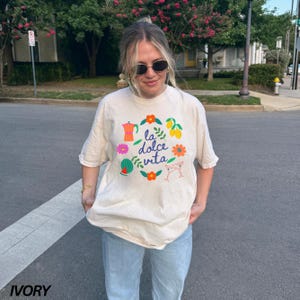 Pu&ograve; includere: Una donna indossa una maglietta beige chiaro con un design grafico colorato che dice "la dolce vita" e un cerchio di limoni, fiori, una caffettiera e bicchieri da martini. La maglietta &egrave; di taglio ampio e ha un collo rotondo.