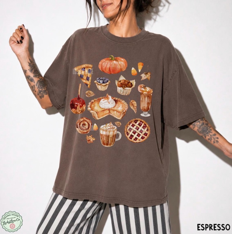 May include: An espresso-colored short-sleeve t-shirt featuring a central graphic print of various autumn-themed treats, including a pumpkin, a slice of pie, a caramel apple, a muffin, a latte, and a lattice-crust pie, all rendered in watercolor style.