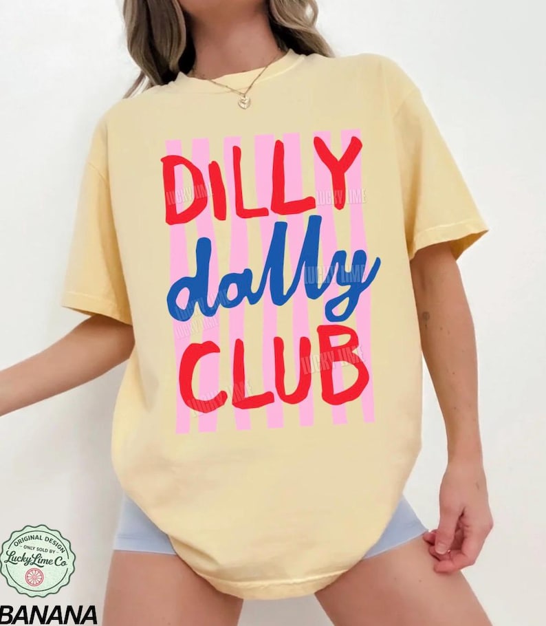May include: A light yellow t-shirt with the words "DILLY dally CLUB" in red and blue lettering, with pink vertical stripes. The shirt has a round neck and short sleeves. The bottom of the shirt is visible.